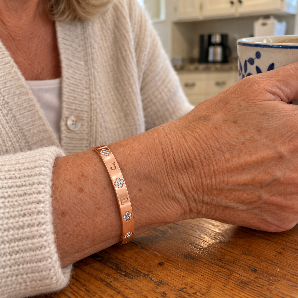 Custom Four-Stone Initial Bangle in rose gold with J / L personalization, worn with a mug at a kitchen table; illustrative phone-style marketing image - Anthology Jewelry