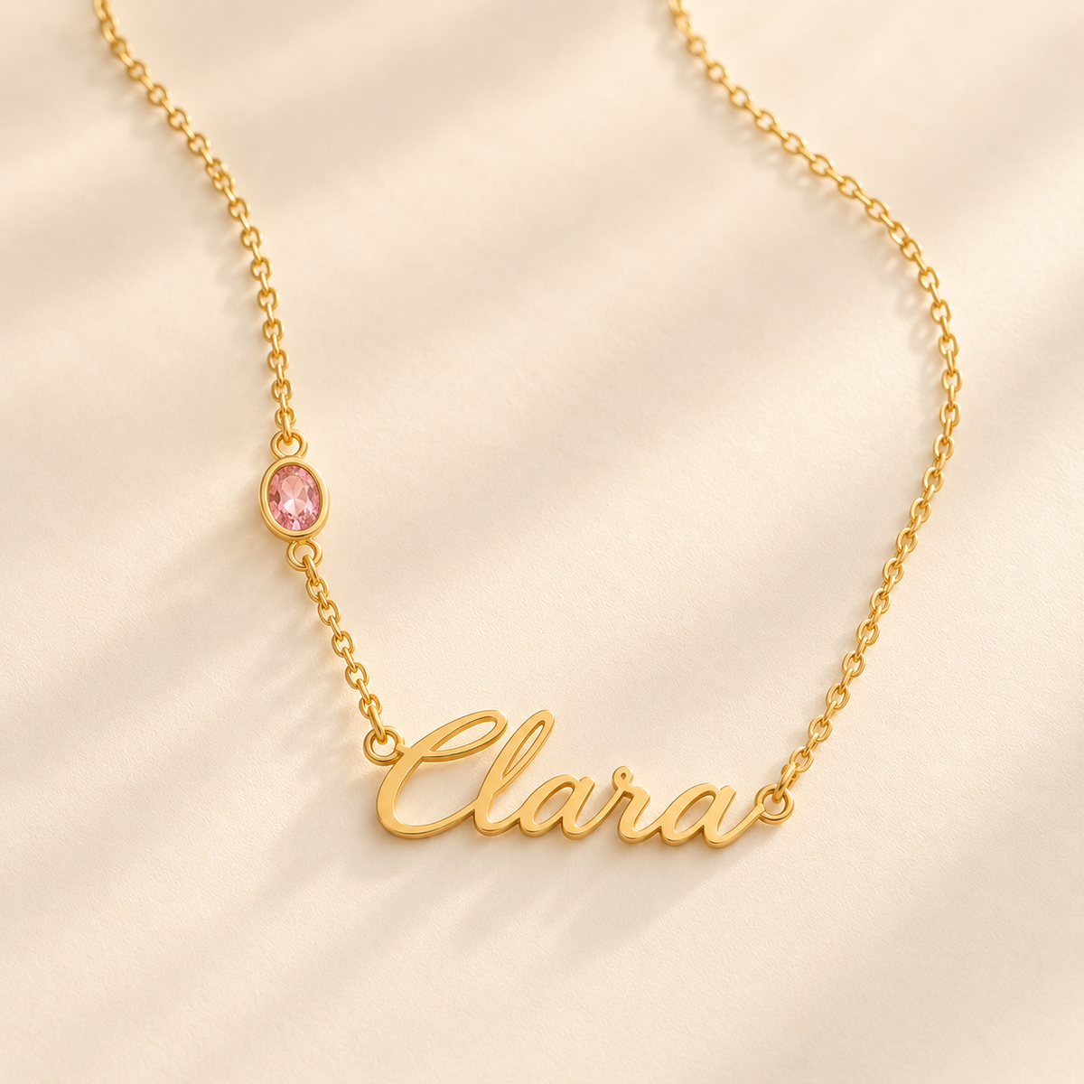 Gold script name and offset birthstone necklace on warm ivory surface