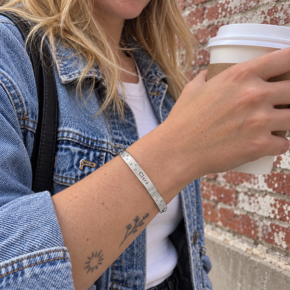 Custom Scattered Stone Name Bangle in silver with Clara personalization, worn holding a takeaway coffee outdoors; illustrative phone-style marketing image - Anthology Jewelry