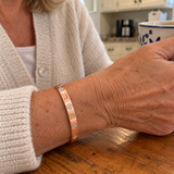 Custom Four-Stone Initial Bangle in rose gold with J / L personalization, worn with a mug at a kitchen table; illustrative phone-style marketing image - Anthology Jewelry