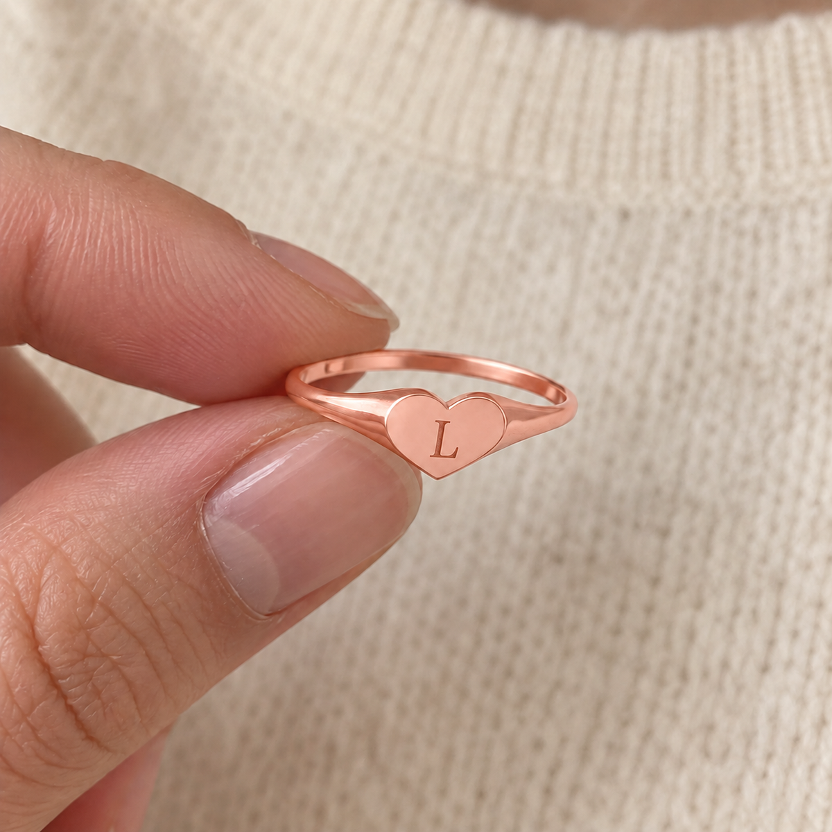 Custom Heart Signet Initial Ring in rose gold with L personalization, casual phone-style view held between fingertips - Anthology Jewelry