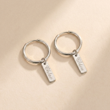 Custom Nameplate Huggie Hoop Earrings hero photo - silver huggie hoop earrings with engraved nameplate charm on cream background Anthology Jewelry
