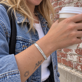 Custom Scattered Stone Name Bangle in silver with Clara personalization, worn holding a takeaway coffee outdoors; illustrative phone-style marketing image - Anthology Jewelry