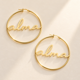 Custom Script Name Hoop Earrings hero photo - gold alma script hoops lying on pale ivory Anthology Jewelry